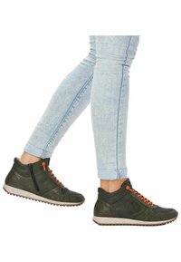 Des baskets montantes vertes avec des lacets orange et une fermeture éclair, présentant des sections perforées et une tige texturée, reposant sur une semelle légère et à motifs.