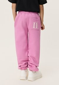 Παιδί που φοράει φαρδιά ροζ φόρμα με πίσω τσέπη που έχει λευκό σχέδιο "II", συνδυασμένο με λευκά αθλητικά παπούτσια και μαύρο μπλουζάκι.