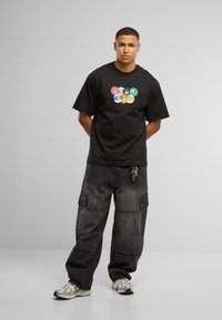 Camiseta de algodón negra con un gráfico colorido de "HOMEBOY", combinada con pantalones cargo de mezclilla oscuros y anchos, y zapatillas reflectantes.
