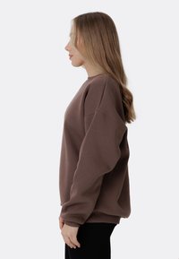 Hnedý oversized sveter s okrúhlym výstrihom, rebrovanými manžetami a voľným strihom, nosený s čiernymi nohavicami. Hladká textúra, minimalistický dizajn.