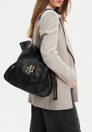 Donna che indossa un blazer beige e pantaloni neri tiene una grande borsa imbottita nera con logo doppia B dorato e tracolla a catena sulla spalla.