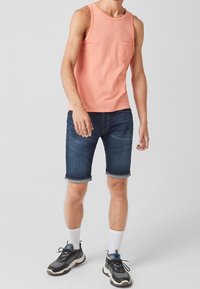 Top de tirantes rosa claro con un bolsillo en el pecho, combinado con shorts de denim azul oscuro que están doblados en el dobladillo. Zapatillas negras con calcetines blancos.