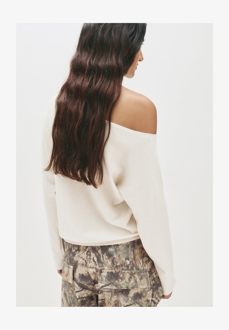 Cremfarbener Off-Shoulder-Pullover mit langen Ärmeln, kombiniert mit gemusterten Cargo-Hosen in Erdtönen und strukturiertem Stoff.