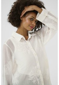 White sheer blouse with embroidered floral patterns, long sleeves, button-down front, and a classic collar. Soft texture, lightweight material.