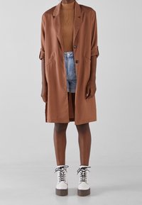 Brązowy długi płaszcz z kołnierzykiem, beżowa prążkowana bluzka, dżinsowe szorty i białe masywne buty na sznurówki. Gładki materiał, luźny krój.