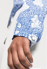 Camicia a fiori blu con sfondo bianco, dotata di polsini con bottoni e un accento metallico circolare, realizzata in tessuto testurizzato.