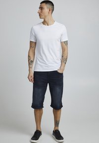 T-shirt en coton blanc à manches courtes, associé à un short en denim foncé arrivant aux genoux et des baskets noires. Présence de tatouages visibles sur les bras.