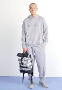 Felpa grigia chiara con logo, pantaloni della tuta grigi abbinati con strisce laterali e uno zaino a camouflage bianco e nero in mano.