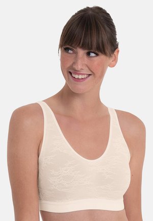 Frau mit braunen Haaren und Pony, die lächelt, trägt ein cremefarbenes ärmelloses Spitzen-BH-Oberteil vor einem schlichten hellen Hintergrund.