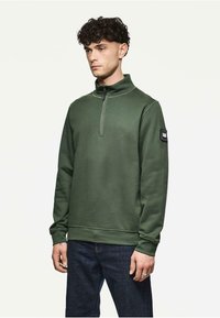 Oliwkowa bluza z długim rękawem z zamkiem na szyi, z ribbowanymi mankietami, o gładkiej fakturze i małą naszywką z logiem na lewym rękawie.