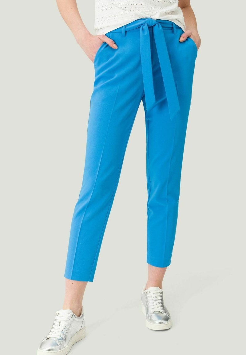 Personne portant un pantalon cropped bleu vif avec une ceinture à la taille et des baskets argentées, les mains calmement dans les poches, montré de la taille vers le bas.