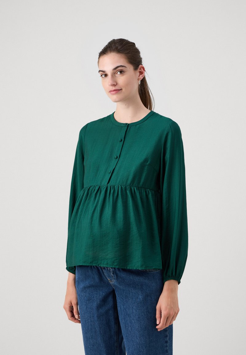 Blouse verte à manches longues en tissu texturé avec un col rond, un devant boutonné et un détail de taille froncée, associé à un jean bleu.