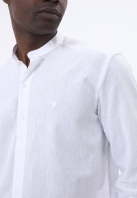 Camisa blanca de lino con cuello mandarín, cierre frontal de botones, textura sutil y un pequeño logo de pájaro bordado en el pecho.