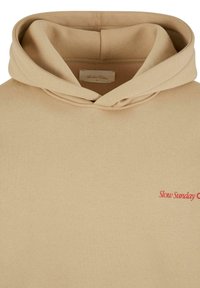 Beige hoodie med en frontkængurulomme og hætte med snor. Har rød tekst "Slow Sunday" på venstre side. Blød stofstruktur.