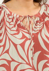 Nahaufnahme einer rot-weiß gemusterten Bluse mit gerafftem Ausschnitt und einer gebundenen Schleife auf der Brust.