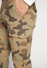 Pantalones cargo en un patrón de camuflaje marrón, verde y beige, con una tela texturizada, que cuentan con un bolsillo lateral y detalles de costura visibles.