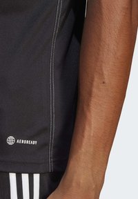 Svart idrottströja med vita söm detaljer och "AEROREADY" logotyp på vänster fåll, visad tillsammans med en texturerad arm.