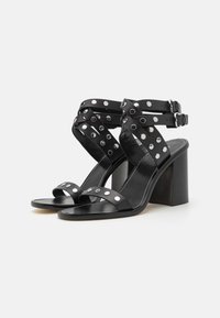 Sandalias de tacón de cuero negro con correas cruzadas, tachuelas circulares plateadas y hebillas ajustables. Cuentan con un tacón cuadrado y un diseño de punta abierta.