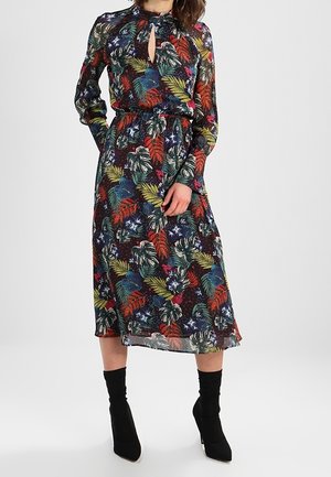 Vrouw draagt een midi-jurk met lange mouwen met kleurrijke bloemen- en bladmotieven, gecombineerd met zwarte enkellaarsjes met hak.