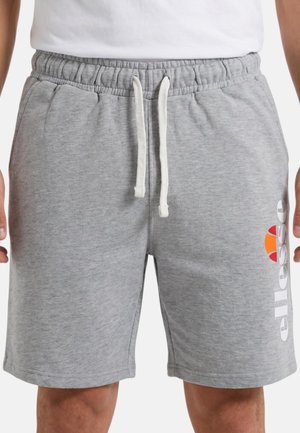 Homme portant un short gris clair à cordon et un t-shirt blanc, avec le logo "ellesse" et un motif orange sur la jambe gauche. Mains le long du corps.