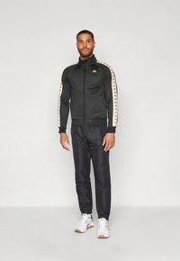 Črna športna jakna s sprednjim zadrgo, ob strani z belimi in zlatimi črtami. Kombinirana s črnimi hlačami in belimi supergami, kar ustvarja športen videz.