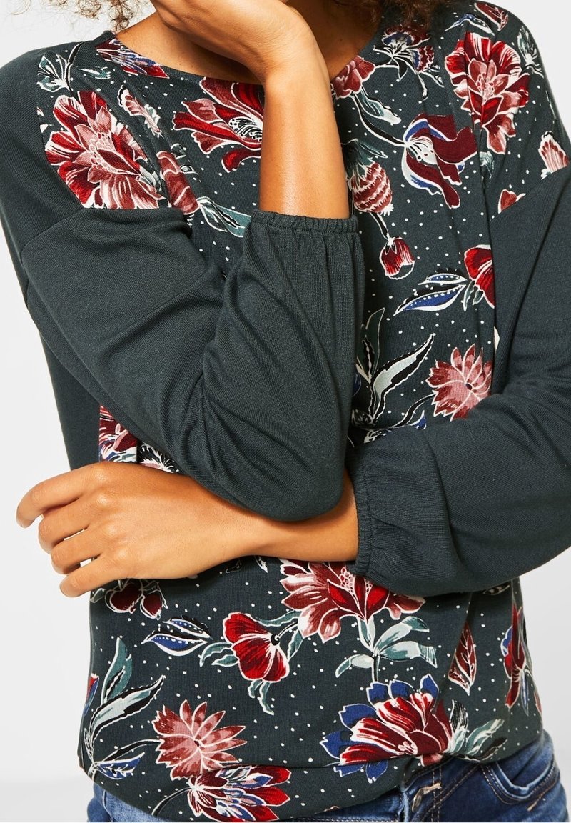 Floral long-sleeve top featuring a dark green body, patterned red and blue flowers, and gathered sleeves, made from soft cotton material.