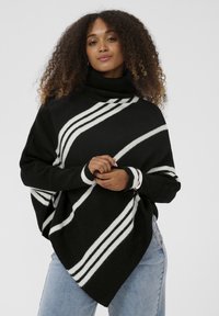 Moteris su garbanojais plaukais, dėvinti juodą golfą su baltais įstrižainiais raštais ir šviesiai mėlynomis džinsomis, stoja su suspaustomis rankomis.