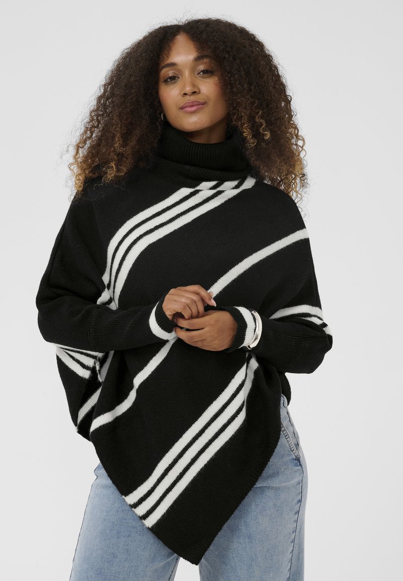 Moteris su garbanojais plaukais, dėvinti juodą golfą su baltais įstrižainiais raštais ir šviesiai mėlynomis džinsomis, stoja su suspaustomis rankomis.