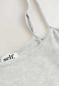 Sivi rebrasti top bez rukava s širokim izrezom, tankim podesivim trakama i etiketom koja na izrezu glasi "self.". Tekstura tkanine.