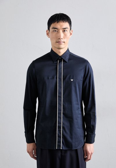 Marineblaues Langarmshirt mit schmaler Passform, das eine kontrastierende weiße Knopfleiste und ein kleines Logo auf der Brust aufweist. Glatte Textur.
