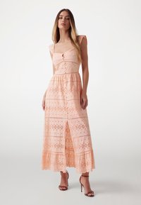 Light peach maxi dress with floral lace texture, short sleeves, button detail at the bust, and a front slit. Features a tiered hem.
