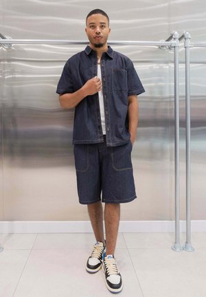 Chemise en denim marine à manches courtes, short assorti et débardeur blanc. Porté avec des baskets noires et blanches dotées d'accents bleus. Fond métallique lisse.