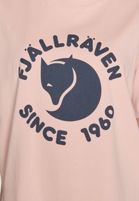 Gaiši rozā kokvilnas T-krekls ar tumši zilu grafiku, kurā attēlots stilizēts lapsa un teksts "FJÄLLRÄVEN SINCE 1960" treknajos burtos.
