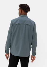 Camicetta a maniche lunghe in tessuto blu verde opaco, con colletto a camicia, dettaglio della yoke posteriore e polsini regolabili. Texture liscia e leggera.