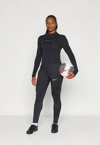 Fekete sportos öltözék, hosszú ujjú felsővel és mintázott, testhez simuló leggingsszel. Egy fehér, fekete részletekkel díszített focilabdát tart a kezében.