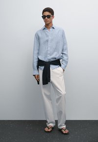 Striped blue and white button-up shirt, white trousers, black sweater tied around waist, sunglasses, and brown flip-flops.