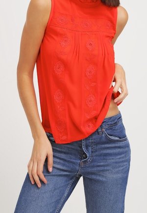 Czerwona, bezrękawnikowa bluzka z kwiatowym haftem, noszona z niebieskimi dżinsami, osoba lekko podciąga bluzkę, odsłaniając talię.