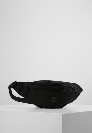 Sort stof bæltetaske med to lynlåslommer og justerbar rem, placeret på en hvid piedestal mod en grå baggrund.