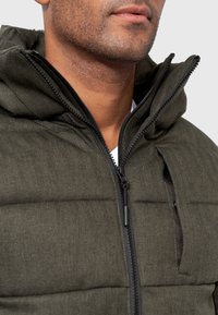 Dunkelgrüne Steppjacke mit Fischgratmuster, ausgestattet mit einem Reißverschluss und einer Kapuze. Texturiertes Material mit horizontalen Abschnitten.