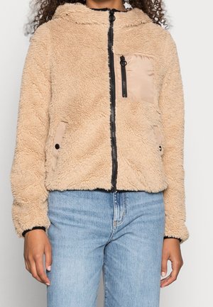 Beige, pluizige jas met rits en capuchon, contrasterende rits en borstzak, gedragen met lichtblauwe spijkerbroek.