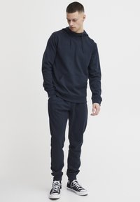 Felpa blu navy con cappuccio e pantaloni abbinati in tessuto morbido, dotati di tasca frontale, cappuccio con coulisse e polsini a coste. Sneakers nere.