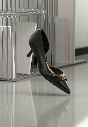 Chaussure noire à talon kitten à bout pointu avec un emblème doré sur l’orteil, présentée en suspension dans l’air devant une surface réfléchissante et une étagère métallique.