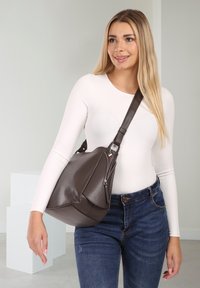 Frau trägt ein weißes Langarmoberteil und blaue Jeans und hält eine große dunkelbraune Leder-Umhängetasche mit einem Reißverschlussdetail.