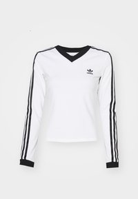Biała koszulka z długim rękawem w serek z bawełny, z czarnymi poziomymi paskami wzdłuż rękawów i czarnym kołnierzykiem. Czarny logotyp Adidas na piersi.