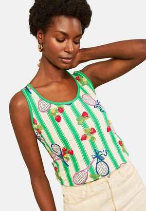 Vrouw draagt een groen-wit gestreepte tanktop met aardbei- en tennisracketpatronen, gecombineerd met lichtgekleurde broek.