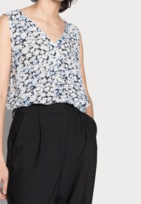 Donna che indossa una blusa floreale senza maniche infilata in pantaloni neri a vita alta con pieghe, con le mani in tasca, mostrata dalla gola ai fianchi.