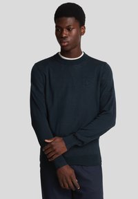 Pullover blu scuro con collo rotondo, maniche lunghe e polsini a coste. Presenta un logo discreto sul petto. Texture liscia, design aderente.