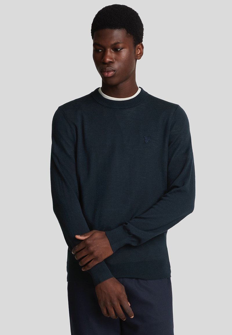 Pullover blu scuro con collo rotondo, maniche lunghe e polsini a coste. Presenta un logo discreto sul petto. Texture liscia, design aderente.