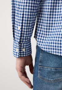 Blå og hvit rutete skjorte med knappet mansjett, kombinert med lyseblå denimjeans. Nærbilde av ermet og hånd plassert nær livet.