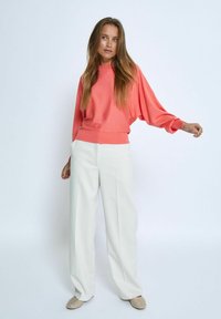 Korallfärgad oversized tröja med sänkt axel och ribbat nederkant, tillsammans med högmidjade vida vita byxor och beigea skopar.